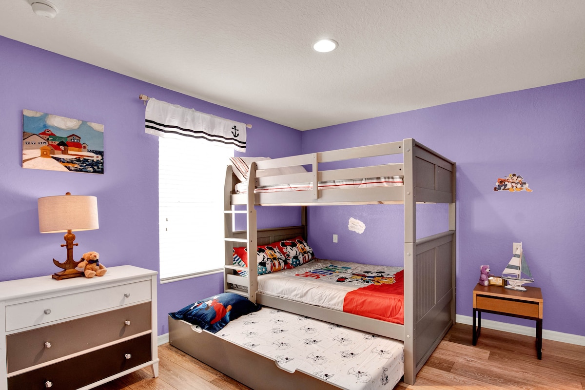 A children's bedroom features purple walls and white trim. A sturdy bunk bed holds colorful bedding, with a lower bed tucked beneath. A small table and lamp provide additional lighting, while nautical-themed decorations and wall art enhance the cheerful environment.