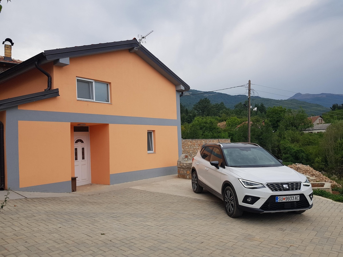 A modern house with a warm orange exterior is shown, featuring a light-colored front door and a spacious driveway. A silver SUV is parked nearby, with a hilly landscape visible in the background under a cloudy sky.
