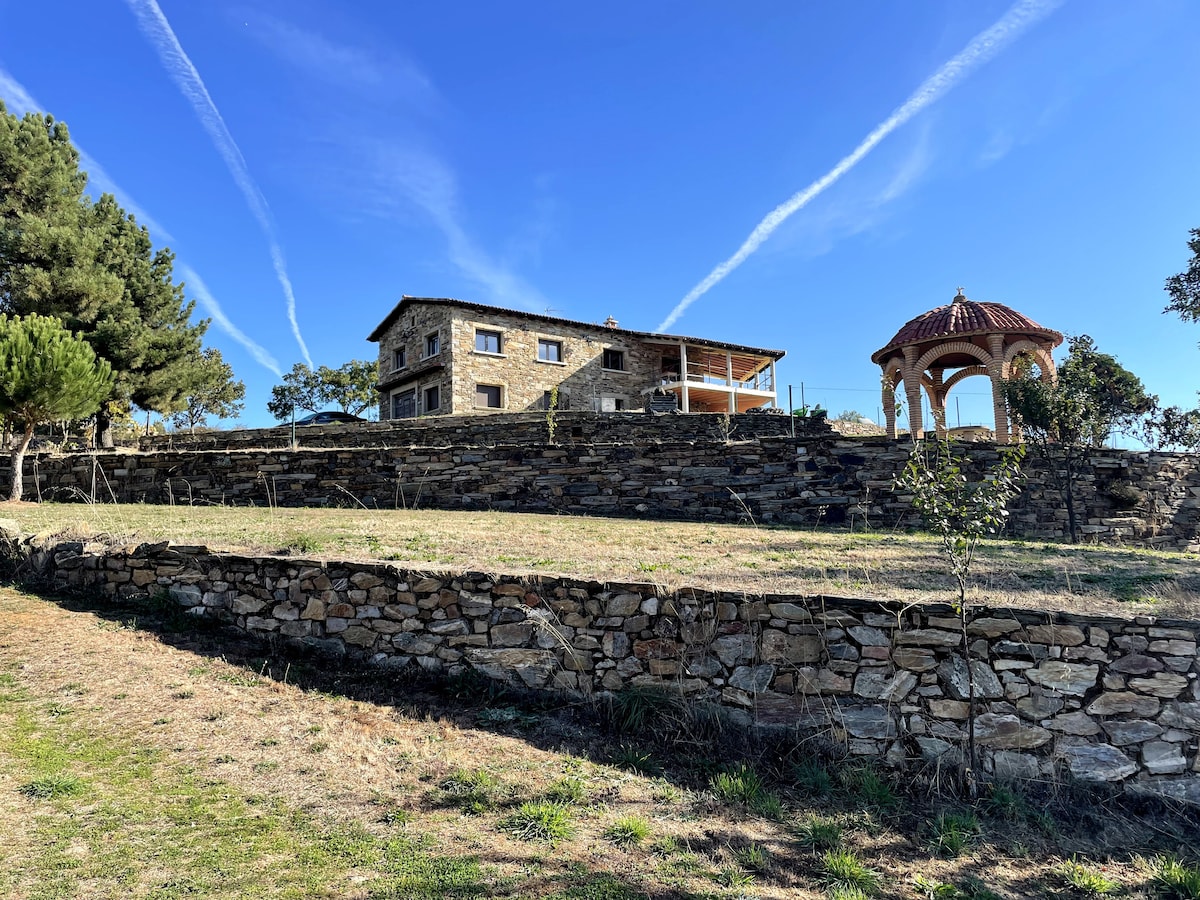 A rustic stone house is situated on a grassy hillside, featuring a covered gazebo nearby. The blue sky is accented by wispy clouds, while tree line adds to the natural surroundings. The elevated placement provides a panoramic view of the landscape.