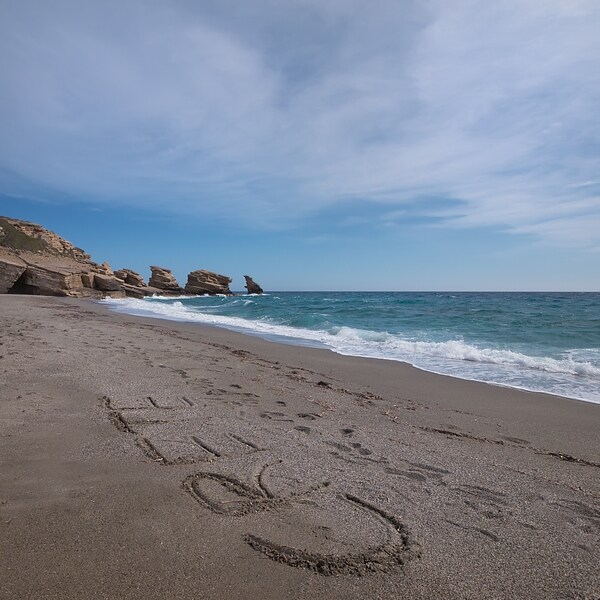 A sandy beach stretches along a clear blue sea, with gentle waves lapping at the shore. Distinct rock formations rise from the water in the distance, while textures in the sand create a natural contrast against the ocean backdrop.