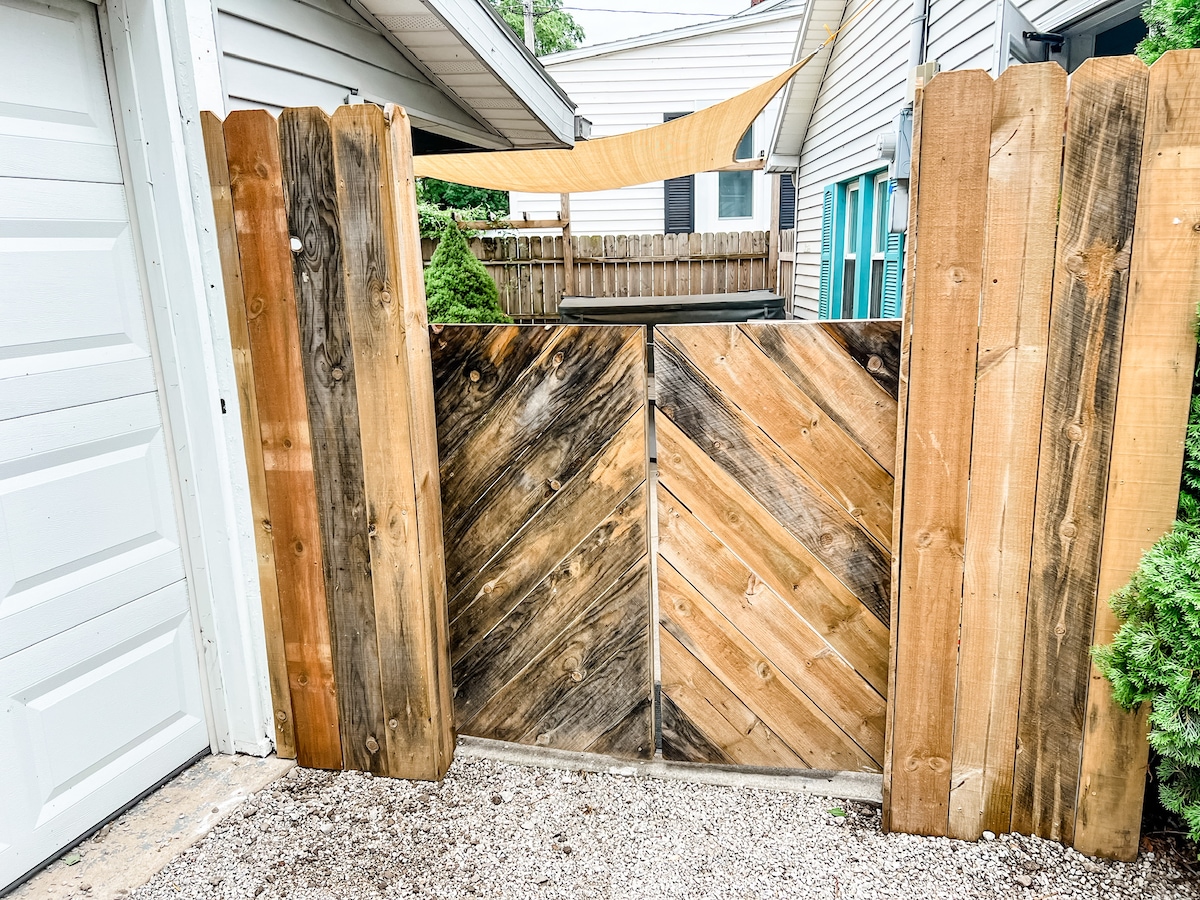 A wooden gate with a chevron pattern stands between two fences, leading into the backyard. The surrounding area features gravel flooring and greenery, providing a welcoming entry point to the outdoor space.