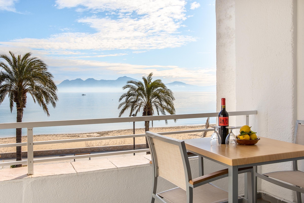 A simple outdoor seating area is visible, featuring a wooden table set for two. A bottle of wine and a bowl of fruit are placed on the table. The view includes calm waters, distant mountains, and palm trees, creating a serene coastal atmosphere.