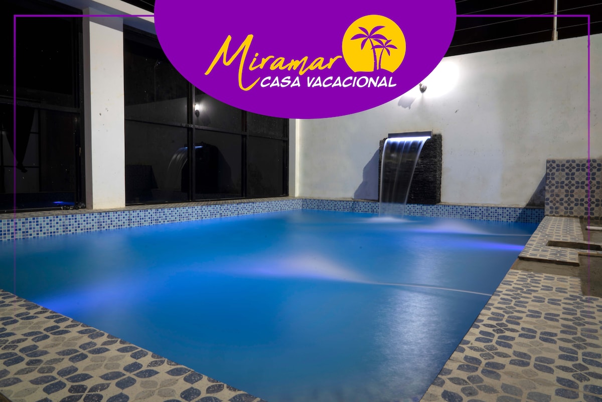 A tranquil swimming pool is featured, illuminated with soft lighting. A waterfall feature adds a serene touch, while the pool is surrounded by smooth stone tiles. Large windows provide a view of the outdoors, creating an open and airy feel.
