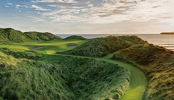 A scenic golf course is framed by gentle hills and lush greenery, with the vast ocean visible in the distance. The sun casts soft light across the landscape, creating a serene outlook over the fairways and mounds, inviting relaxation and enjoyment of the natural surroundings.