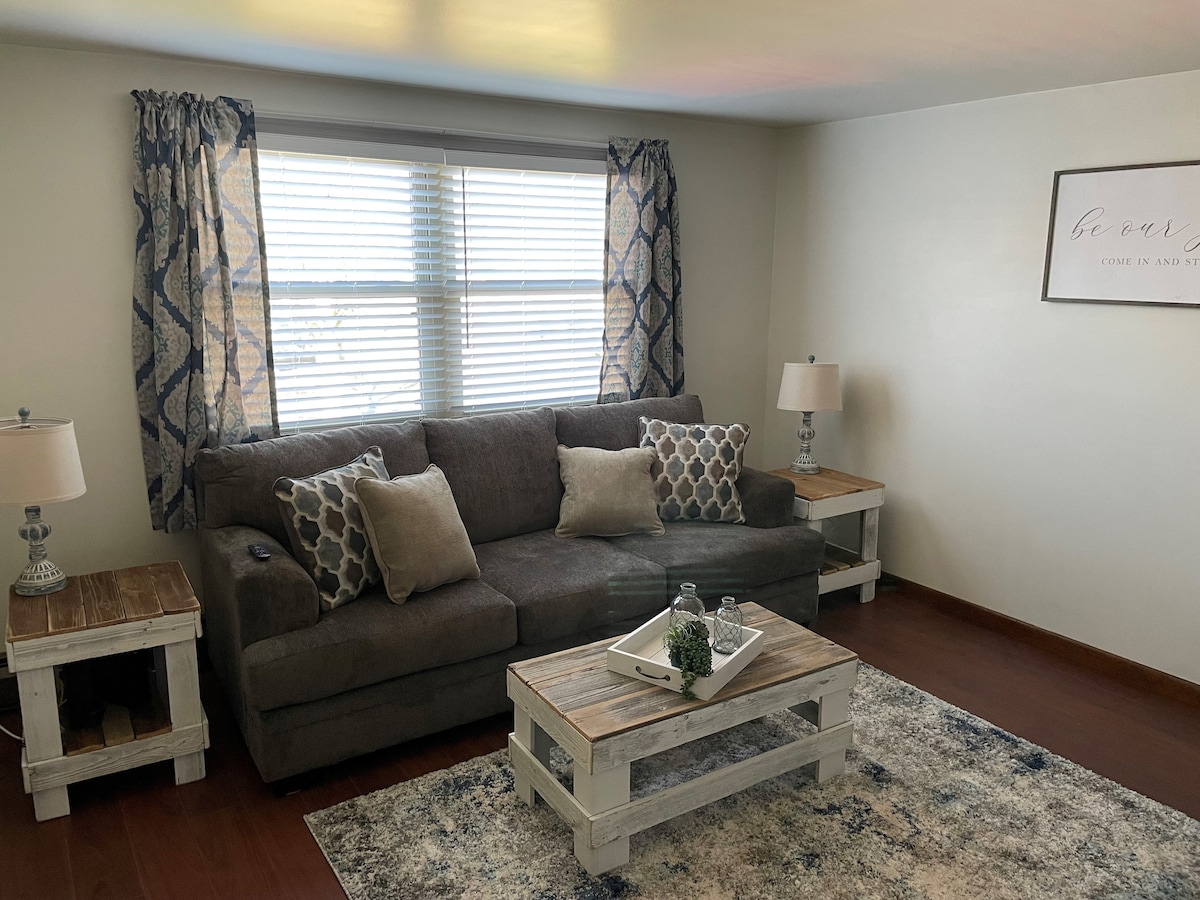 A comfortable living area is designed with a neutral-toned sofa adorned with decorative pillows. Two wooden side tables with lamps flank the sofa, and a stylish area rug anchors the space. Natural light streams through a window, enhanced by patterned curtains.