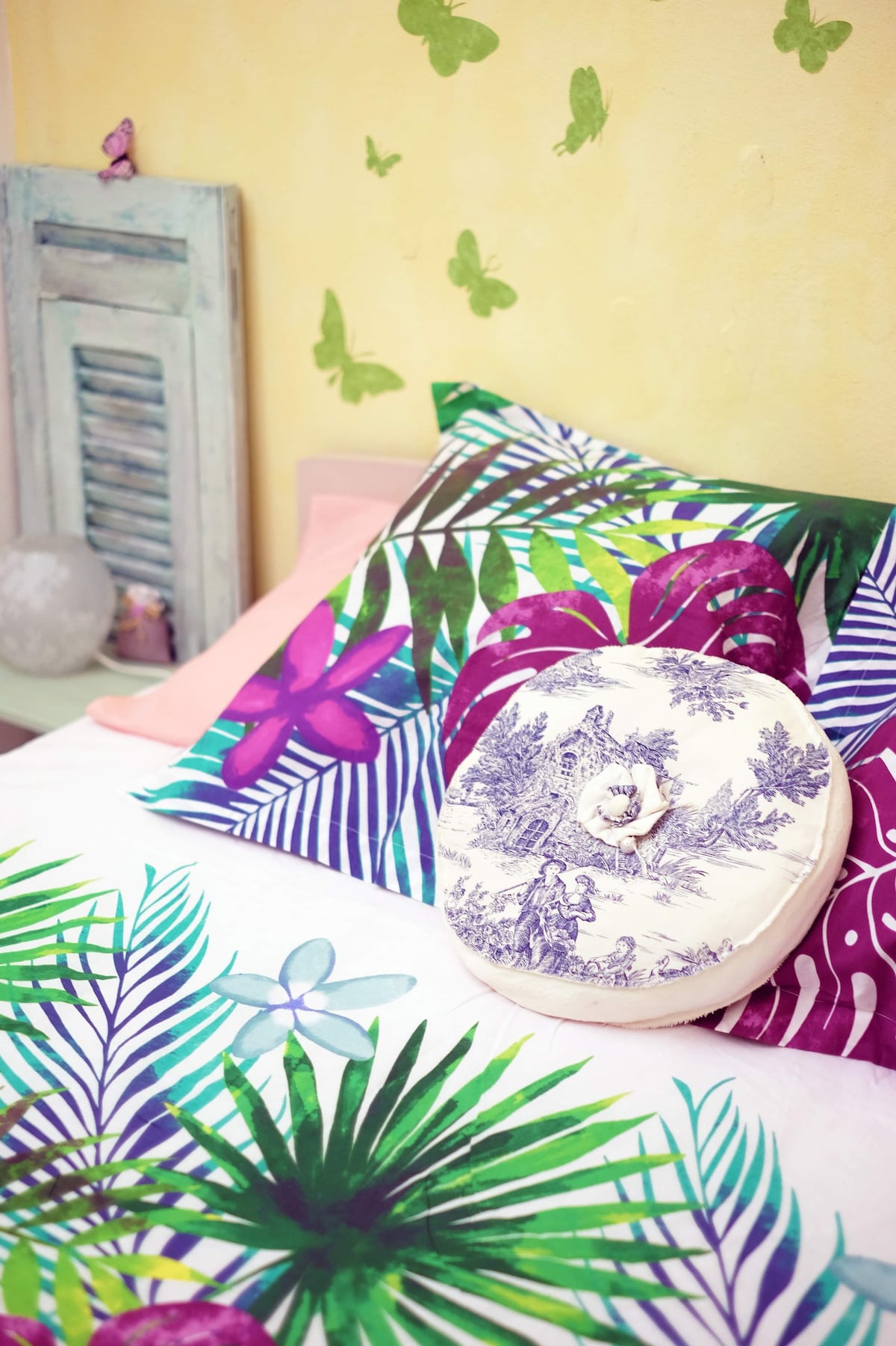 A vibrant bed is adorned with patterned bedding featuring tropical foliage. Decorative pillows in various colors and designs add interest, while a round cushion with a vintage illustration serves as a stylish accent. Soft yellow walls and butterfly accents enhance the cheerful ambiance.