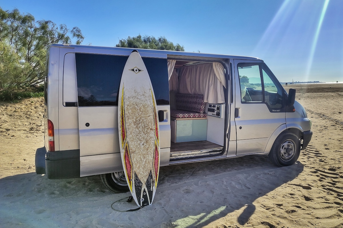 A van with an open sliding door is parked on sandy terrain. A surfboard leans against the exterior, highlighting a casual beach vibe. The interior features curtains and patterned upholstery, providing a comfortable atmosphere for relaxation and travel.