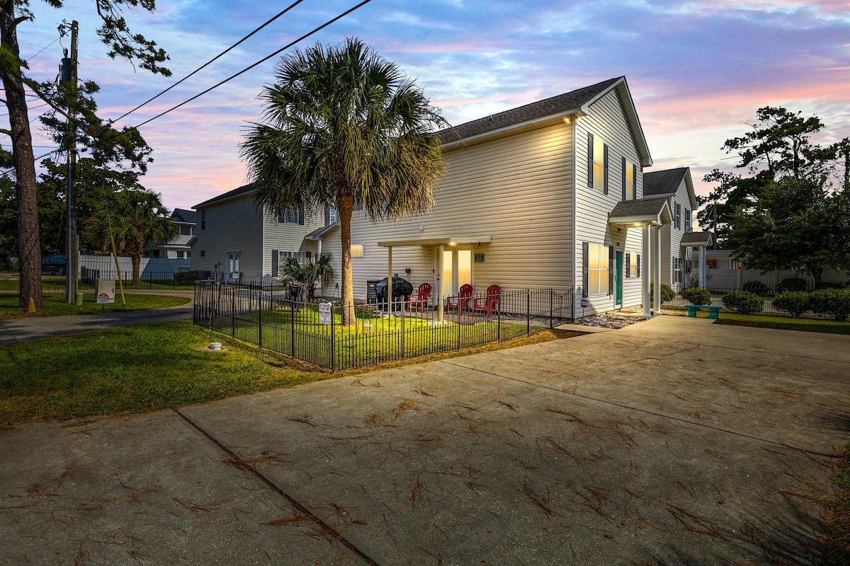 The exterior of the beach home features a well-maintained yard with a small, fenced patio area and vibrant green grass. Adirondack chairs are arranged for relaxation. A spacious driveway is visible, framed by palm trees and surrounding greenery, offering a welcoming entrance.