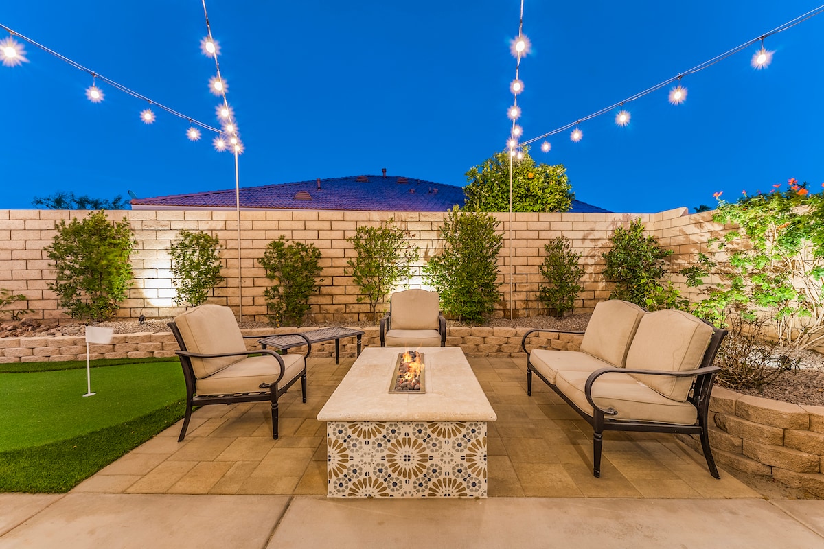 An outdoor seating area features cream-colored lounge chairs surrounding a decorative fire pit. String lights hang above, illuminating the space. Decorative plantings provide greenery against a stone wall backdrop, while a putting green is visible in the corner.