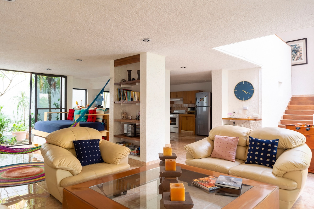 A spacious living area features light-colored furniture arranged around a coffee table. Natural light filters through large windows, revealing a glimpse of the garden. A staircase leads to the upper level, and an organized kitchen is visible in the background.