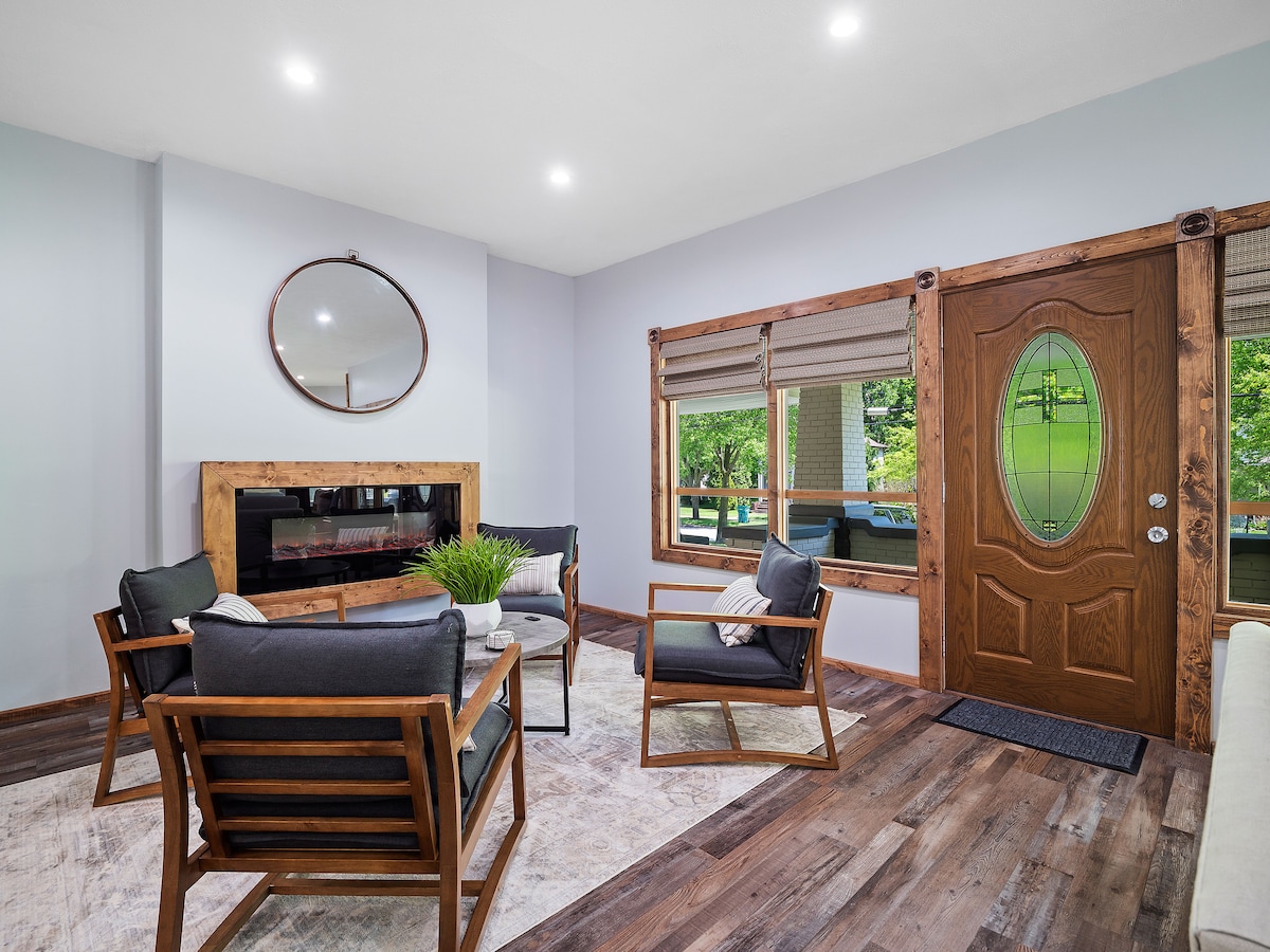 The living area features a modern design with comfortable seating arranged around a central electronic fireplace. Large windows provide natural light, while a wooden door adds warmth. A mirror above the fireplace reflects the welcoming ambiance of the space.