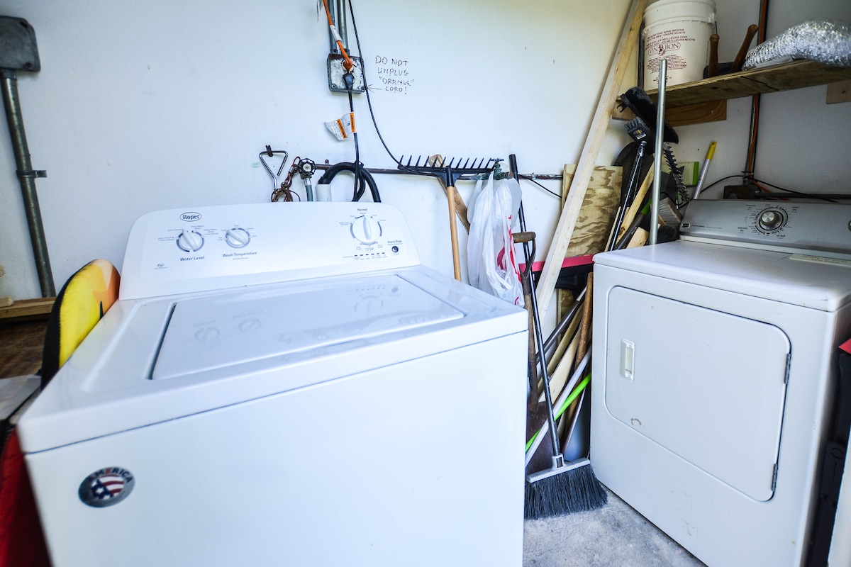 A laundry area is equipped with a washing machine and a dryer. Various cleaning supplies and tools, including a broom and rake, are neatly organized on shelves and the floor, offering a functional space for laundry needs.