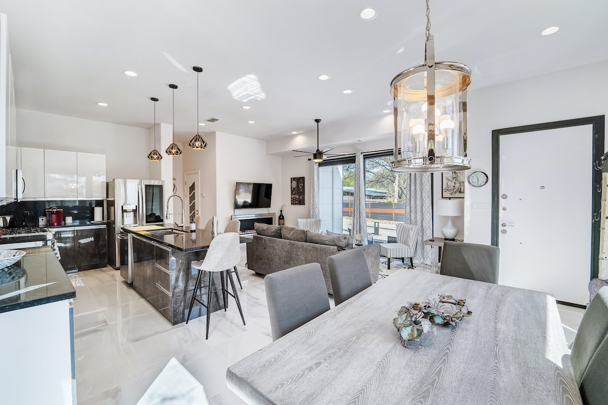 An open concept living space showcases a modern kitchen featuring a large island with seating, sleek cabinetry, and a commercial range. The dining area features a wooden table with seating for six, and the living room is furnished with gray sofas. Large sliding glass doors invite natural light and provide access to an outdoor patio.