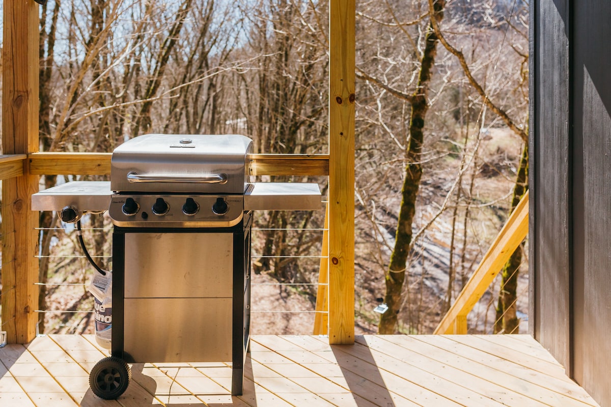 An outdoor grill is positioned on a wooden deck, surrounded by a backdrop of tall trees with bare branches. A glimpse of the river can be seen in the distance, enhancing the natural setting.