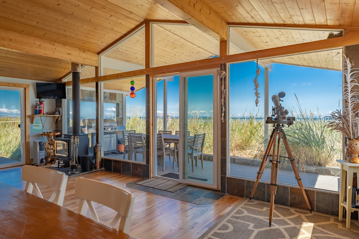 An open-concept living area features large glass sliding doors that provide views of the beach and sea grass outside. A wood stove is positioned next to the dining table, while a telescope stands ready for observing the natural surroundings.