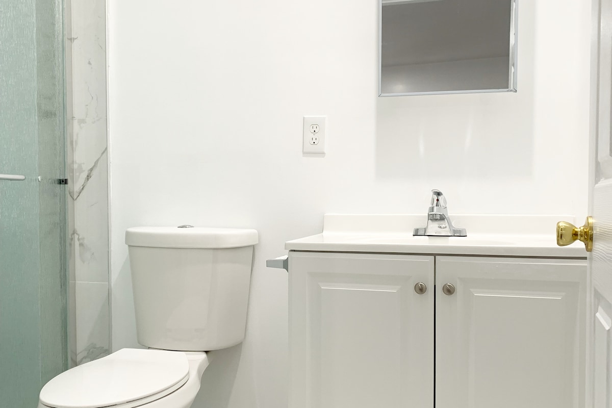 A clean, modern bathroom is featured, showcasing a white toilet and a sleek vanity with storage below. A chrome faucet is seen on the countertop, along with a large wall mirror that reflects the bright, well-lit space. A glass shower enclosure is visible in the corner.