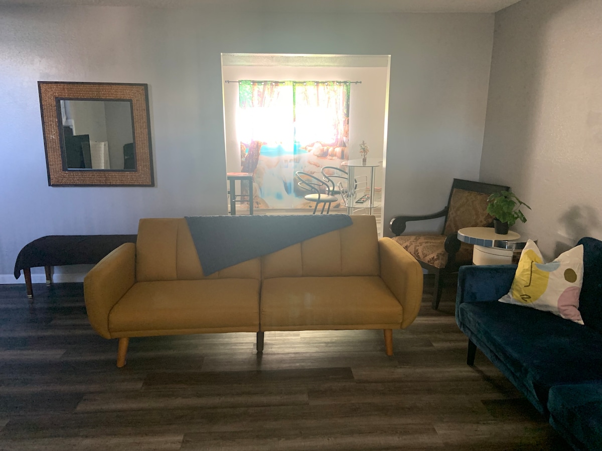 A welcoming living area features a yellow sofa paired with a blue armchair. A small side table holds a plant, and light filters through a window adorned with colorful curtains, illuminating the space. A mirror is mounted on the wall, enhancing the room's bright ambiance.