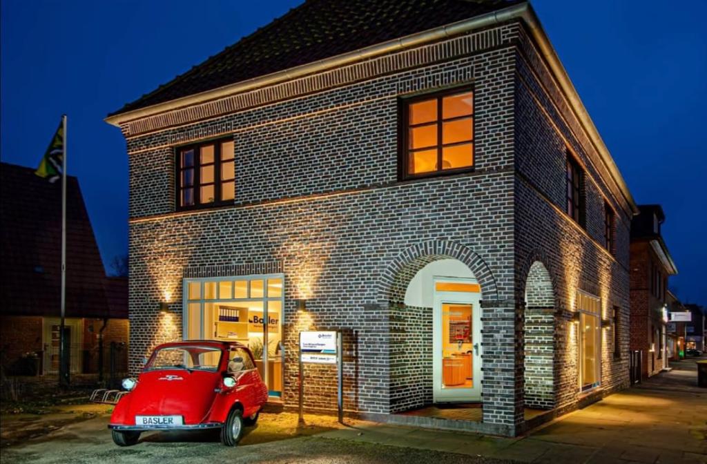 A charming brick building is illuminated in the evening light, featuring large windows and an inviting entrance. A vintage red car is parked in front, adding character to the scene. The structure's architectural details are highlighted, enhancing its unique aesthetic.