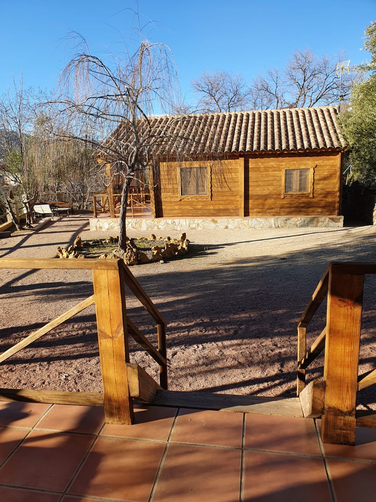 A wooden cabin is situated in a natural setting, featuring a rustic roof and large windows. Surrounding the cabin is a gravel area with a stone fire pit and trees providing shade. A wooden railing leads to the entrance, enhancing accessibility.