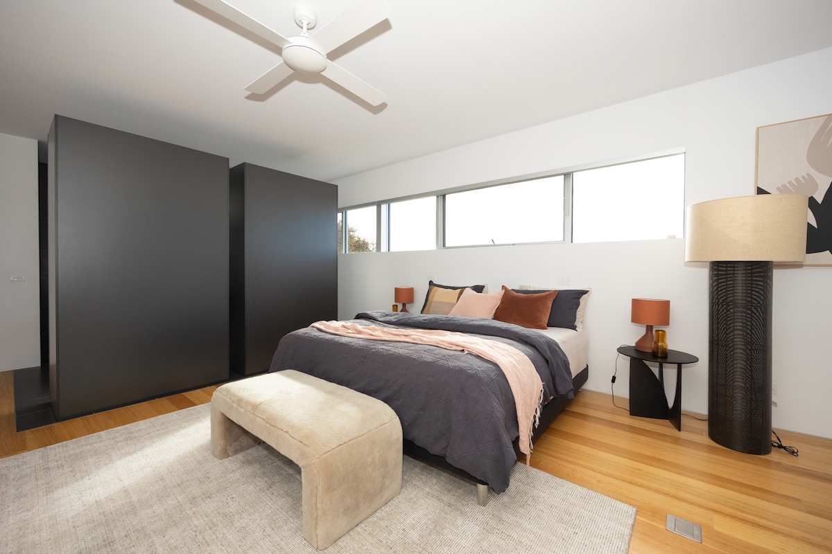 A spacious bedroom features a king bed dressed in dark linens, complemented by accent pillows in warm tones. A bench sits at the foot of the bed, while modern lamps provide ambient lighting. Large windows allow natural light to fill the room, enhancing its open feel.