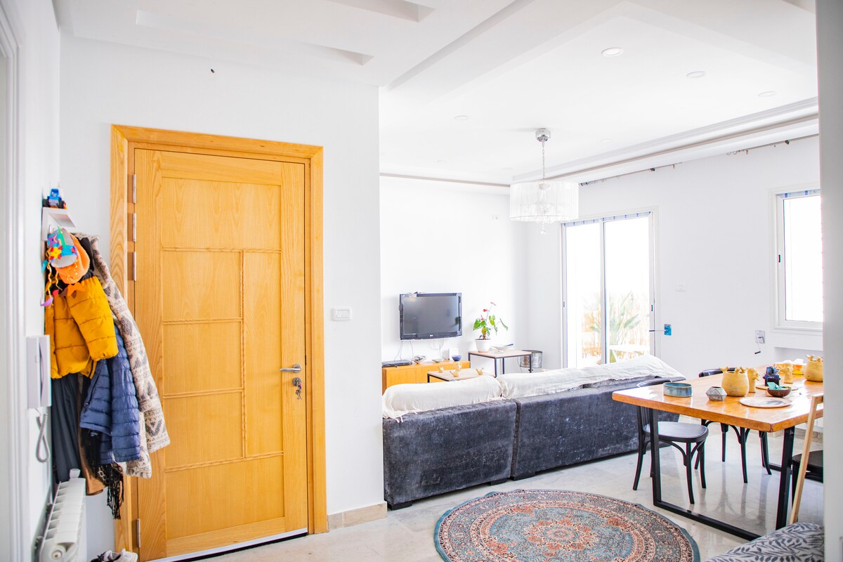 A bright living area showcases a large sofa and a wooden dining table. Natural light enters through the glass doors leading to a balcony. A television is positioned on a cabinet nearby, and a colorful area rug lies on the tiled floor.