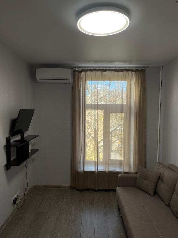 A bright room features a large window with sheer curtains allowing natural light to fill the space. A comfortable beige sofa is positioned against the wall, and a flat-screen TV is mounted on a sleek shelf, adding a modern touch to the decor.
