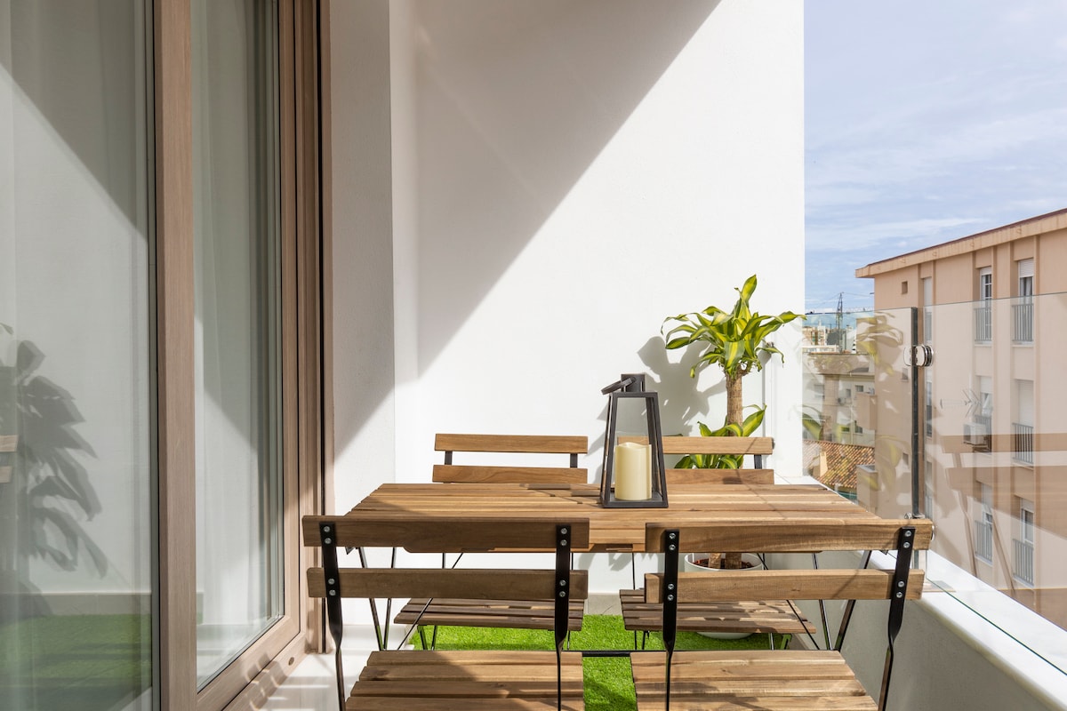 A small balcony features a wooden table and four matching folding chairs, positioned on a green lawn-like surface. A lantern rests on the table, and a potted plant adds a touch of greenery. Natural light illuminates the space, with large glass doors providing access.
