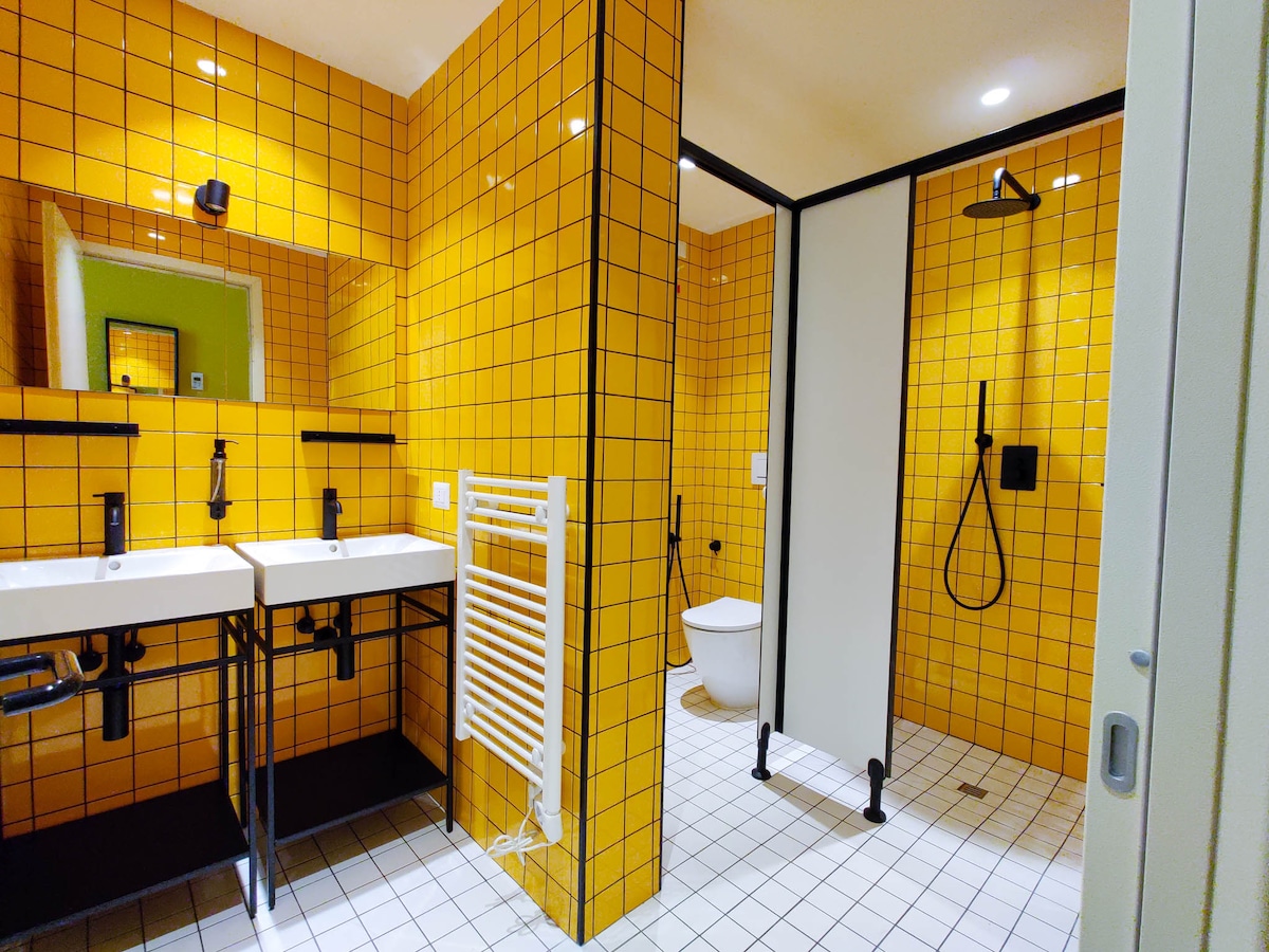 The shared bathroom features vibrant yellow tiled walls and modern fixtures. Two sinks with black fixtures are positioned on a black frame. A spacious shower area, separated by a white partition, includes a handheld showerhead and a toilet. Bright lighting enhances the cheerful atmosphere.