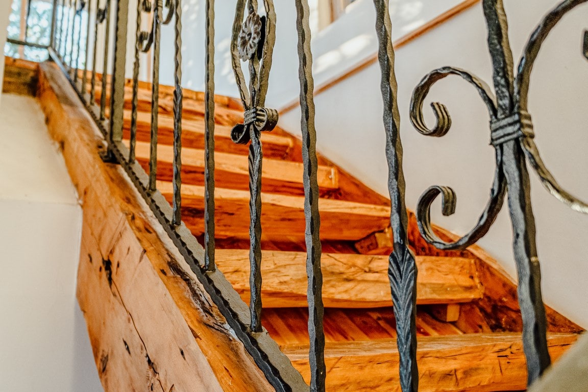 An interior staircase features sturdy wooden steps with a natural finish, complemented by decorative wrought iron railings. Light filters in from nearby windows, highlighting the craftsmanship of the stairway and the inviting atmosphere of the space.
