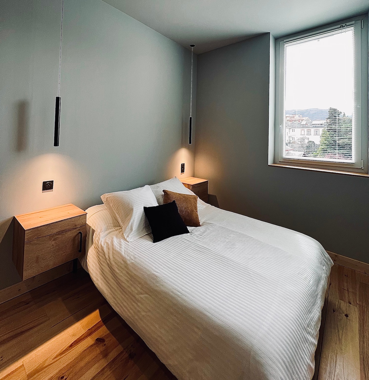 A well-lit sleeping area features a spacious bed with crisp white linens, flanked by minimalistic wooden nightstands. Soft lighting is provided by hanging fixtures, and a window allows natural light to fill the room while offering a view of the surroundings.