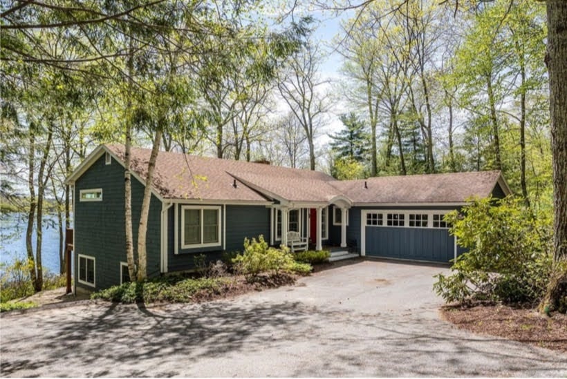 A charming lakefront home is surrounded by lush greenery, featuring a cheerful blue exterior and a welcoming front porch. A driveway leads to a one-car garage, while large windows invite natural light and provide scenic views of the nearby lake.
