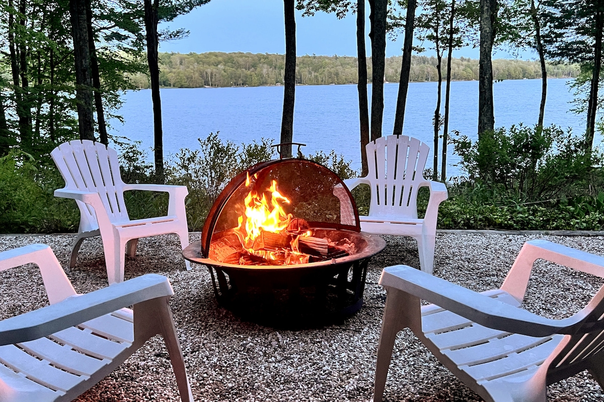 A fire pit surrounded by four white Adirondack chairs is situated on a gravel patio. Flames dance within the fire pit, while a picturesque lake framed by lush green trees is visible in the background, creating a peaceful setting for relaxation.