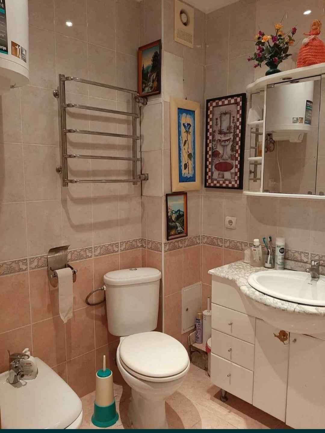 A compact bathroom is equipped with a white toilet and a small sink set into a light-colored vanity. A mirror is mounted above the sink, and a towel rack is attached to the wall. Decorative artwork is displayed alongside floral accents, adding character to the space.