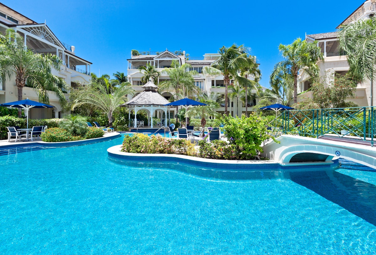 A freeform swimming pool is surrounded by lush greenery and palm trees, with shaded seating areas under umbrellas. The scene includes three residential blocks in the background, set against a clear blue sky, creating a serene environment for relaxation.