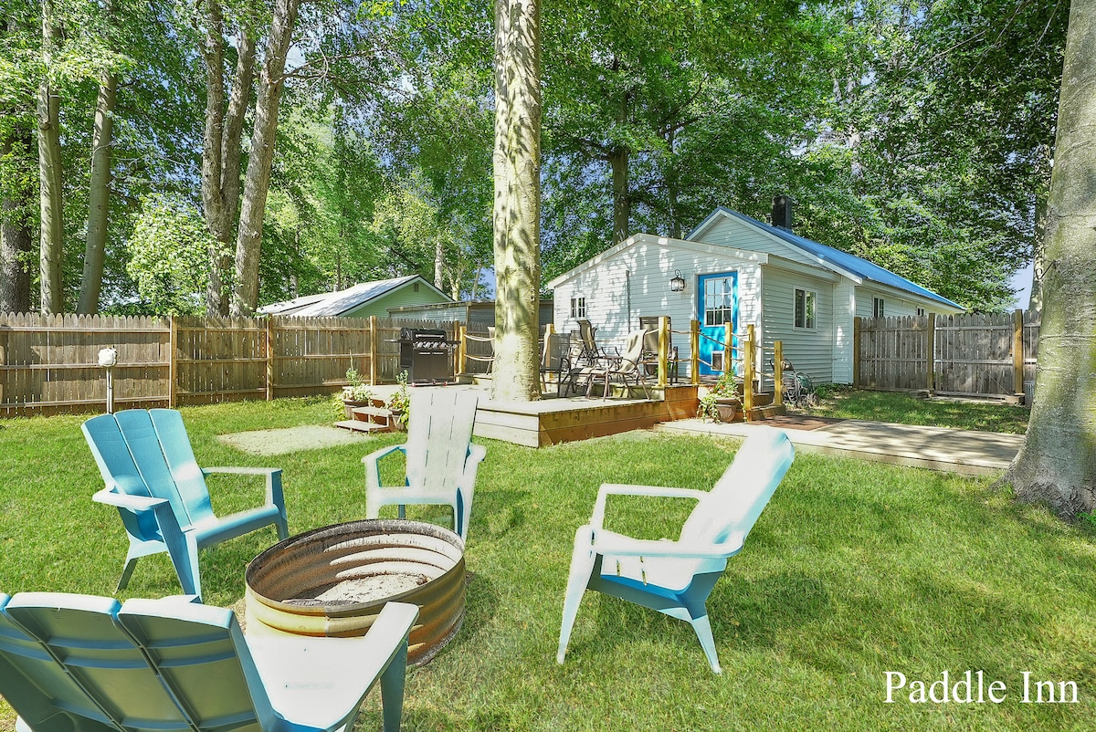 A spacious backyard is featured with green grass and a circular firepit surrounded by several light blue adirondack chairs. A wooden deck with outdoor furniture is visible in the background, along with trees providing shade and a fence marking the property boundary.