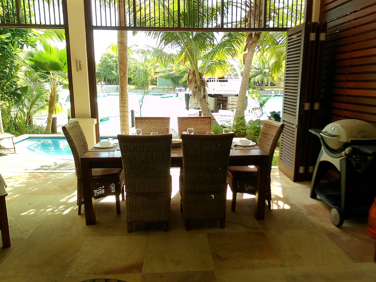 An outdoor dining area is presented with a wooden table surrounded by woven chairs. A grilling station is visible nearby. Lush palm trees frame the view of the sparkling pool, creating a serene environment for meals under natural light.