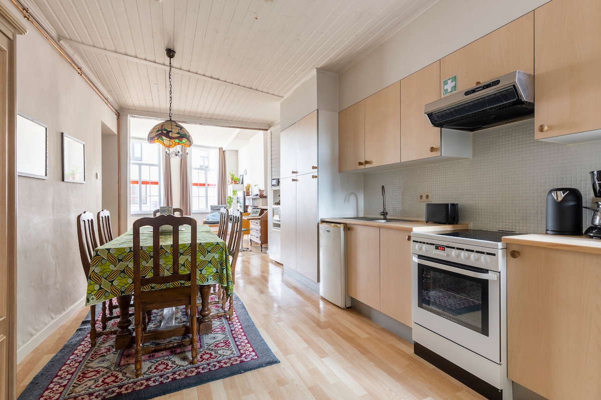A spacious kitchen and dining area features wooden cabinetry and a large dining table surrounded by chairs. Natural light fills the space through large windows, highlighting the intricate tablecloth design. Appliances include an oven, stove, and coffee maker, enhancing the functional layout.