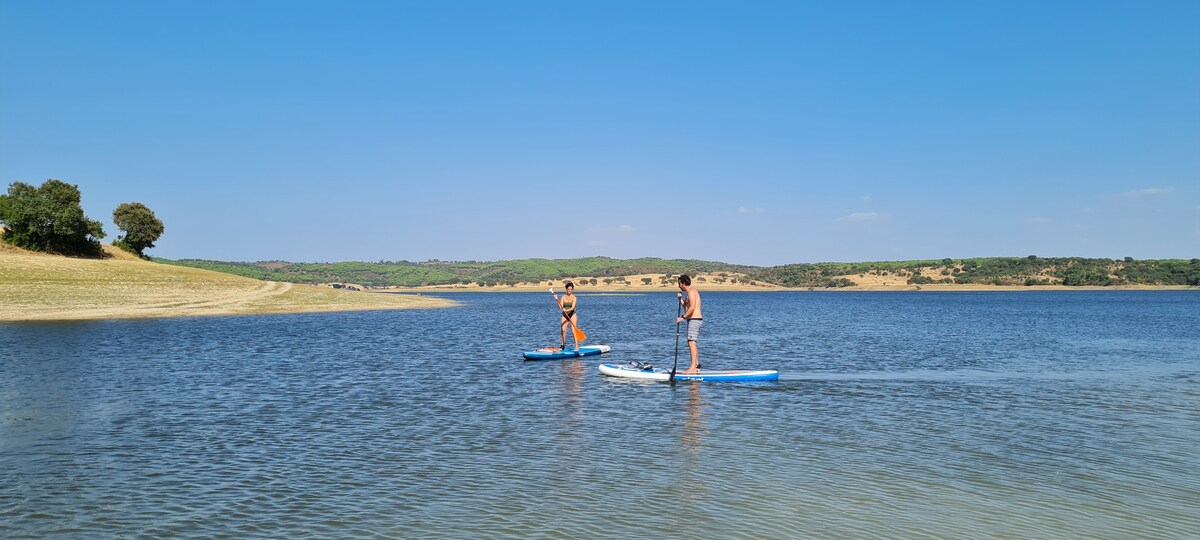 Two individuals are seen paddle boarding on a calm lake, surrounded by gently sloping green hills. The clear blue sky reflects on the water's surface, creating a serene atmosphere.