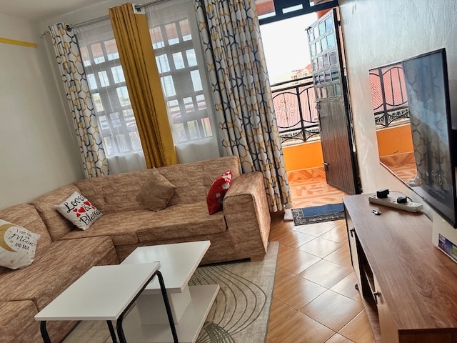 A cozy living area features a large sectional sofa adorned with decorative pillows. Two side tables are positioned in front, complemented by a light rug. Sunlight filters through sheer curtains covering large windows, illuminating the space. A wooden TV stand is placed against the wall.