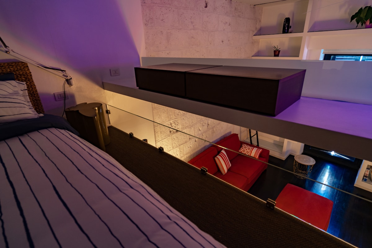 A loft bed is positioned above a cozy sitting area, featuring a red sofa with decorative pillows. Natural light streams in from a window, highlighting the textured stone wall and minimalist white shelving in the background.