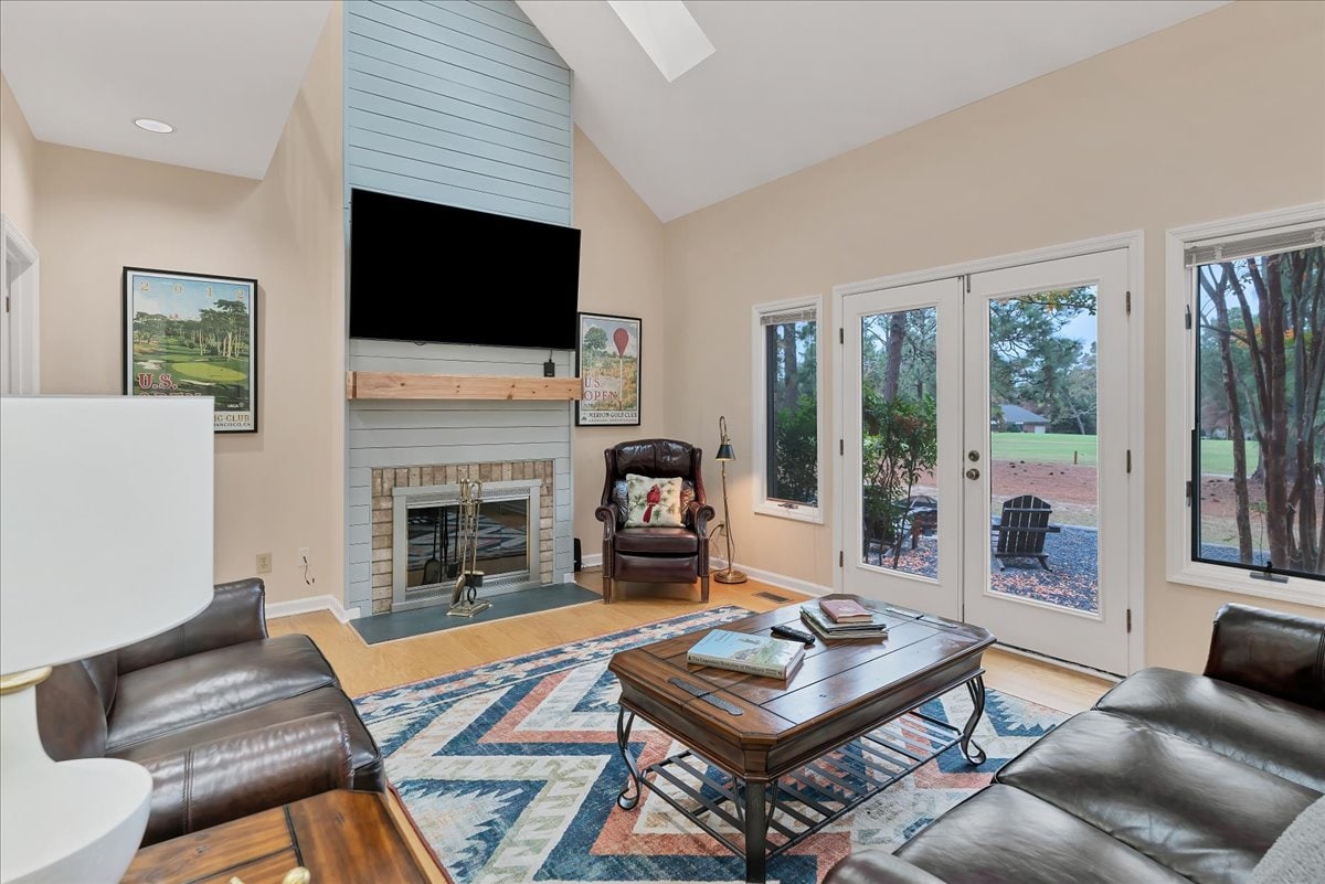 A spacious living area features comfortable leather seating arranged around a central wooden coffee table. A brick fireplace is situated beneath a mounted television, while large windows provide views of the outdoors, enhancing the space with natural light.