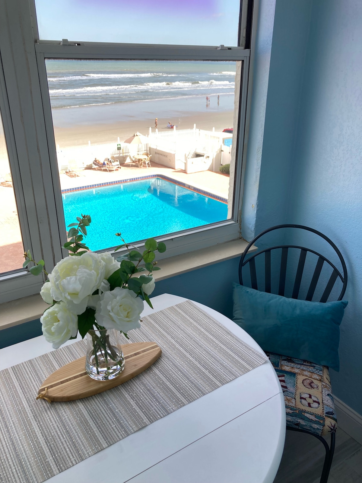 A cozy dining area is positioned by a window, offering views of the pool and beach. A small table is set with a vase of white flowers and a decorative wooden tray. A blue cushion adds comfort to the black chair, enhancing the inviting setting.