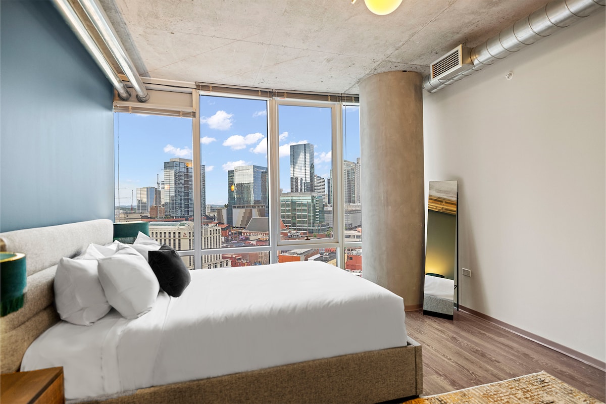 A modern bedroom features a plush queen bed with crisp white linens. Floor-to-ceiling windows provide scenic views of the city skyline, enhancing the natural light in the space. A full-length mirror and minimalist decor contribute to a sleek, contemporary atmosphere.