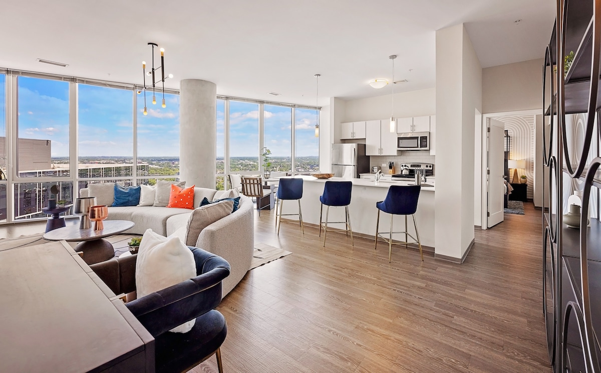 A spacious living area showcases large floor-to-ceiling windows, providing ample natural light. Modern furnishings include a cozy sofa and stylish dining chairs, while the kitchen features contemporary appliances. A view of the surrounding landscape is visible in the background, enhancing the open atmosphere.
