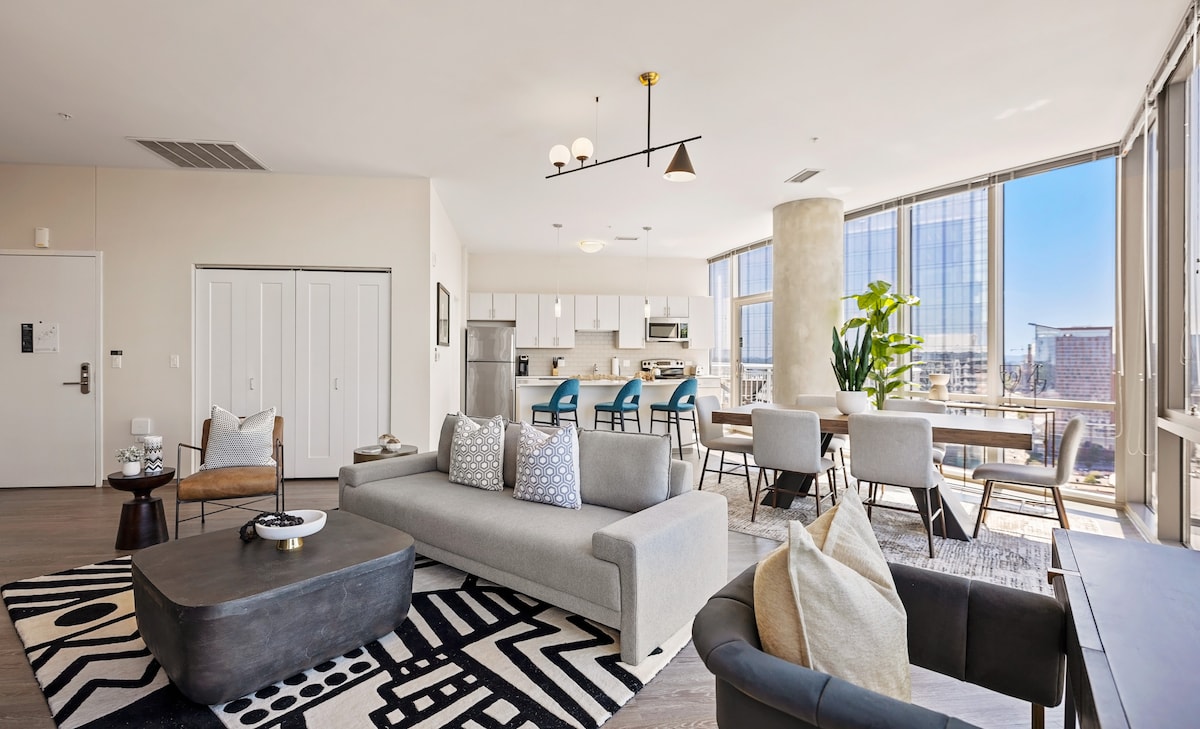 A spacious living area features large floor-to-ceiling windows that invite abundant natural light. A contemporary gray sofa is positioned next to a dark coffee table. A dining area with a wooden table and blue chairs complements the modern kitchen in the background.