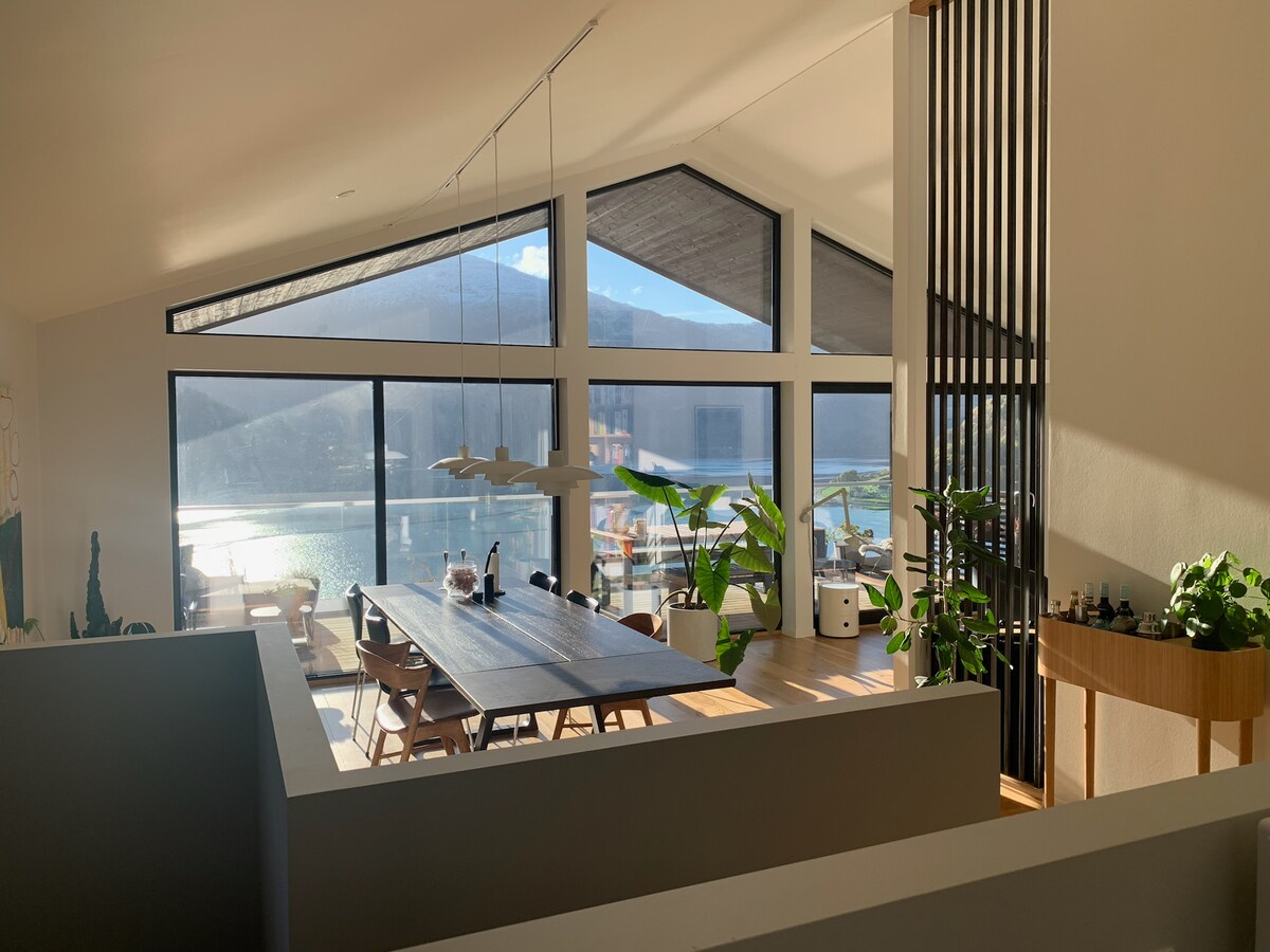 An open living area is highlighted by large windows that allow natural light to fill the space. A long dining table is lined with several wooden chairs, while potted plants are positioned strategically around the room, contributing to the modern interior design.