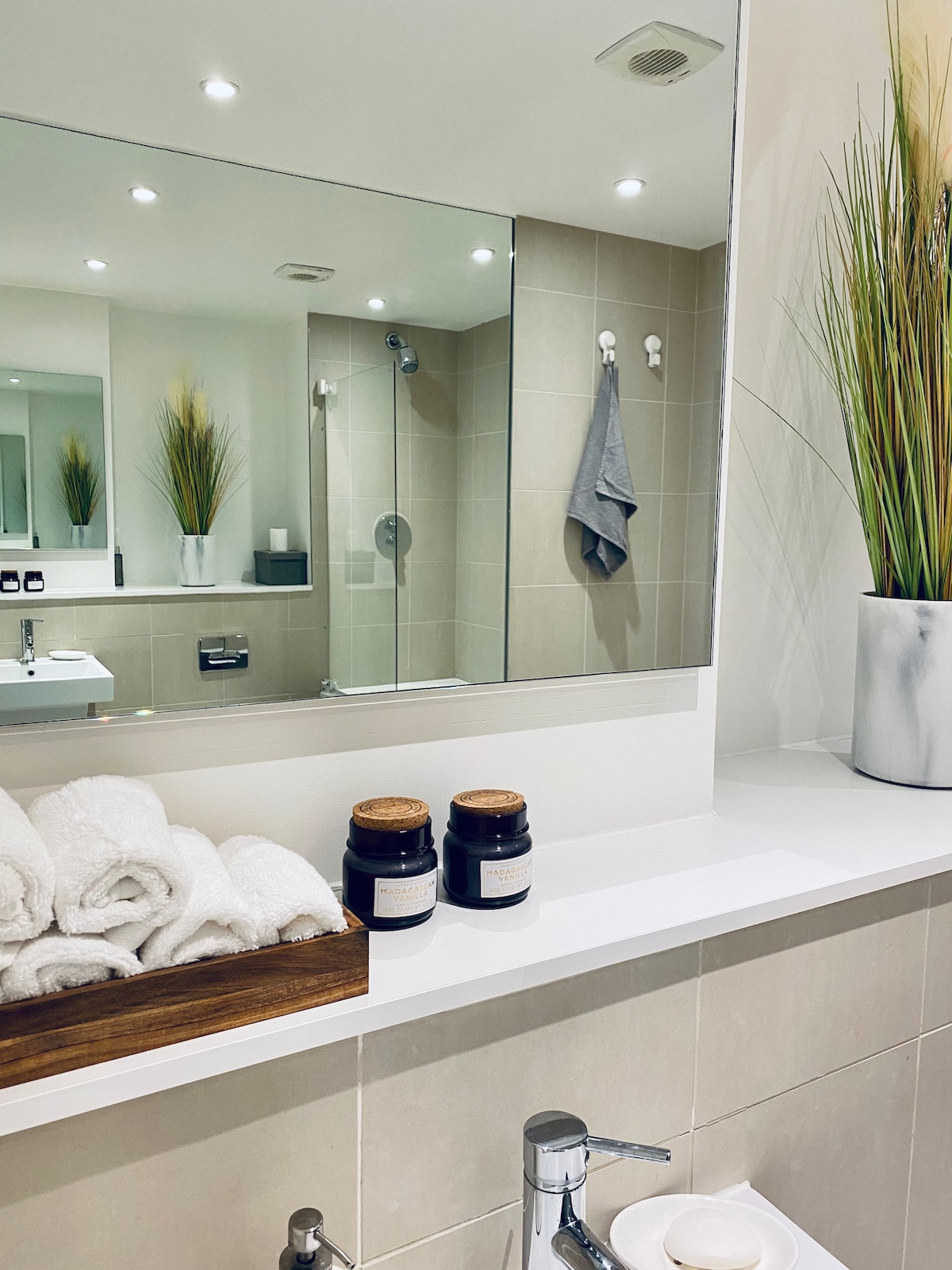 A clean and modern private bathroom is highlighted by a large mirror and soft lighting. Plush towels are neatly arranged on a wooden tray. A shower area is visible in the background, with a plant adding a touch of greenery to the decor.