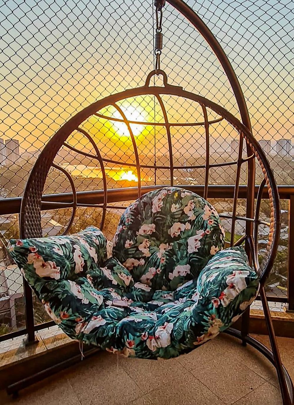 A hanging chair with a circular design is situated on a balcony, featuring a green floral cushion. The background displays a sunset with warm hues reflected on the water, framed by a protective netting and a panoramic city view.