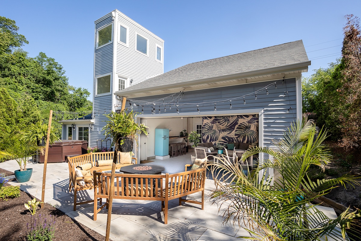 A spacious outdoor patio features a wooden seating area surrounding a fire pit, complemented by tropical-inspired décor. Lush greenery and potted plants are visible, adding to the relaxing ambiance. The cabana's glass overhead door opens to invite the natural surroundings, enhancing the outdoor experience.
