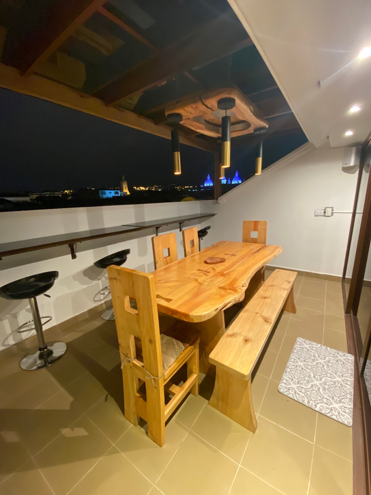 An outdoor balcony is featured, furnished with a wooden dining table and benches, complemented by high bar stools. Soft lighting fixtures hang above, enhancing the evening ambiance, while the cityscape is visible in the background, adding a scenic backdrop to the space.